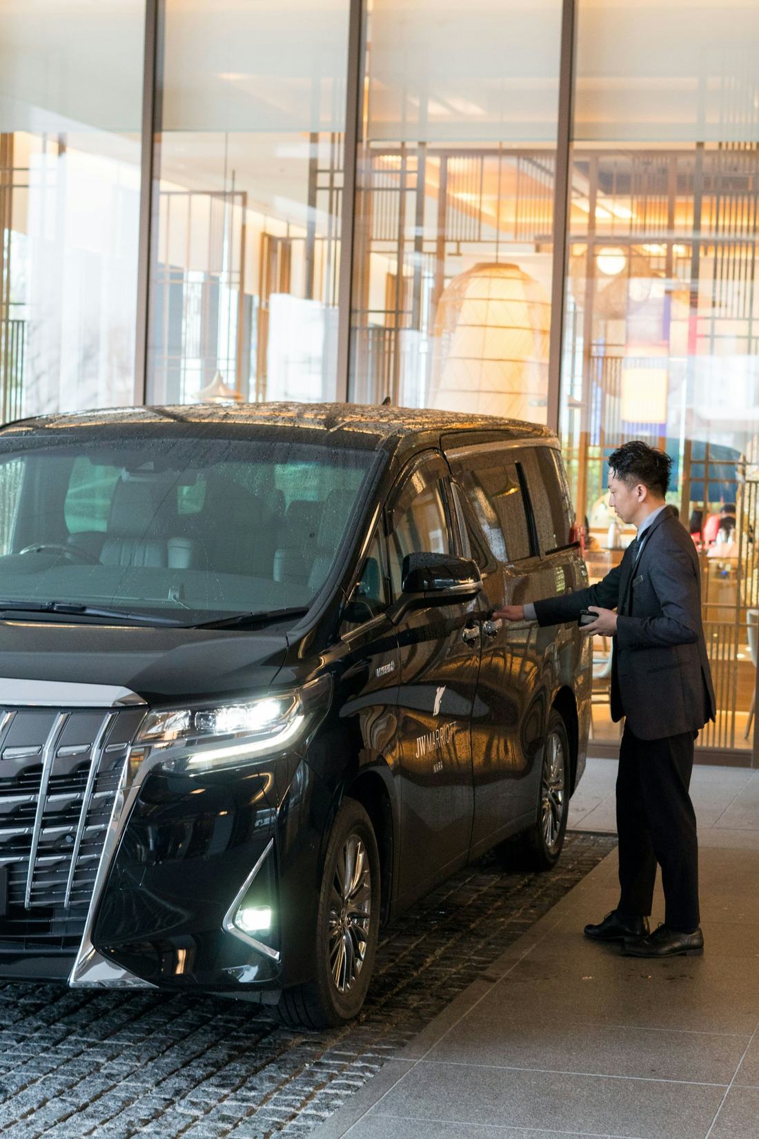 A chauffeur in a dark suit opening the rear door of a black van at a hotel entrance